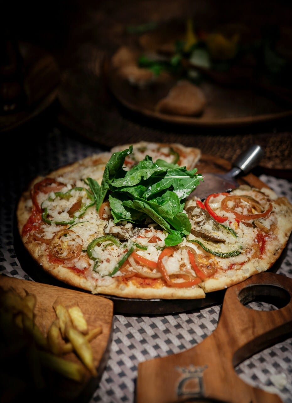 Whole vegan pizza on a wooden board, topped with cheese, sliced peppers, onions, and a pile of fresh rocket in the centre, served with fries on the side.